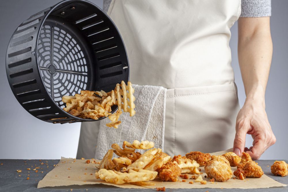 A woman is dumping fresh made potato waffle fries from basket onto a countertop together with chicken nuggets. She fried them in air fryer using very little fat. A healthy homemade convenient snack.