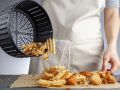 A woman is dumping fresh made potato waffle fries from basket onto a countertop together with chicken nuggets. She fried them in air fryer using very little fat. A healthy homemade convenient snack.