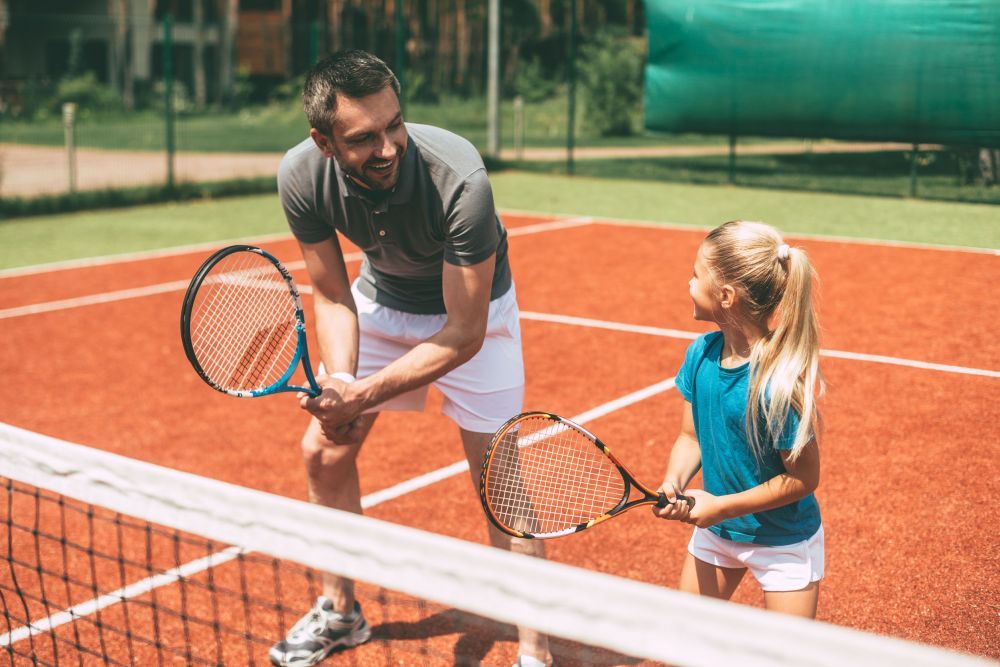 Practicing tennis. Cheerful father in sports clothing teaching his daughter to play tennis while both standing on tennis court