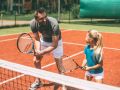 Practicing tennis. Cheerful father in sports clothing teaching his daughter to play tennis while both standing on tennis court